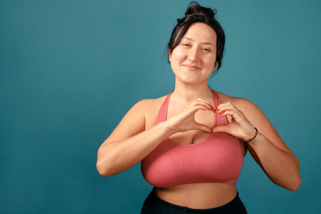 Happy plus size positive woman. Happy body positive concept. I love my body. Attractive overweight woman posing on camera in the studio and make heart from her fingers. Girl is wearing a coral bra