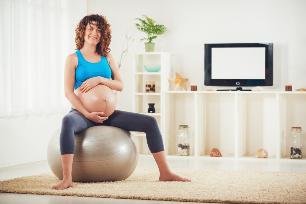 Beautiful young pregnant woman sitting on a fitness ball and relaxing at the home.